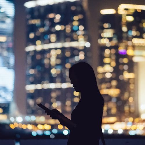 Silhouette of woman using digital tablet device in city at night, with illuminated and blurry commercial skyscrapers in the background.